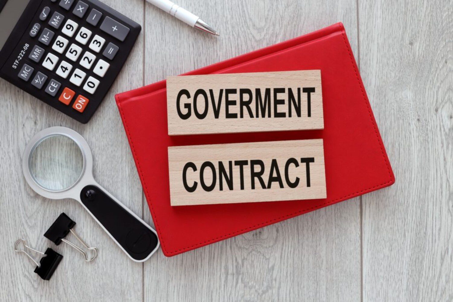 Wooden blocks reading "GOVERNMENT CONTRACT" sit on a red folder, surrounded by a calculator, magnifying glass, and office supplies.