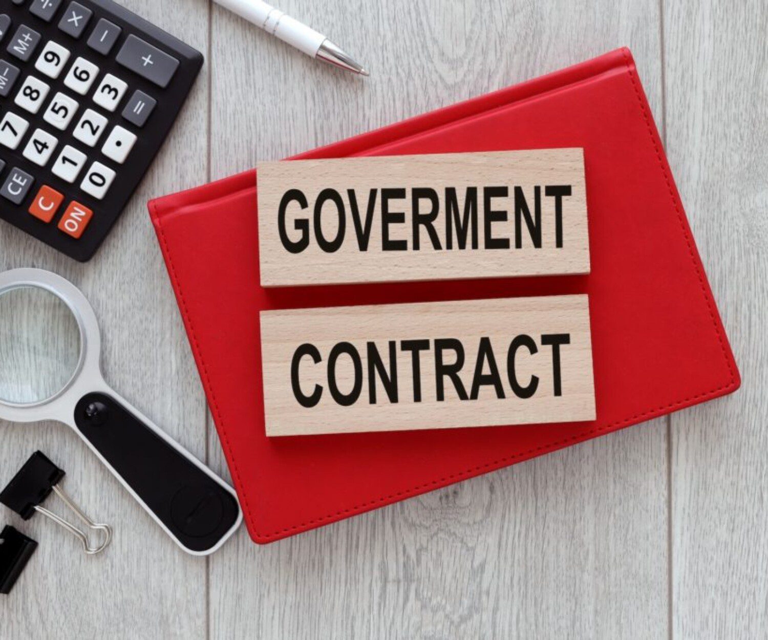 Wooden blocks reading "GOVERNMENT CONTRACT" sit on a red folder, surrounded by a calculator, magnifying glass, and office supplies.