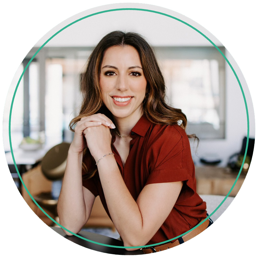 A woman with long brown hair and wearing a red shirt smiles warmly, resting her chin on her hands. She sits in a bright, modern workspace.
