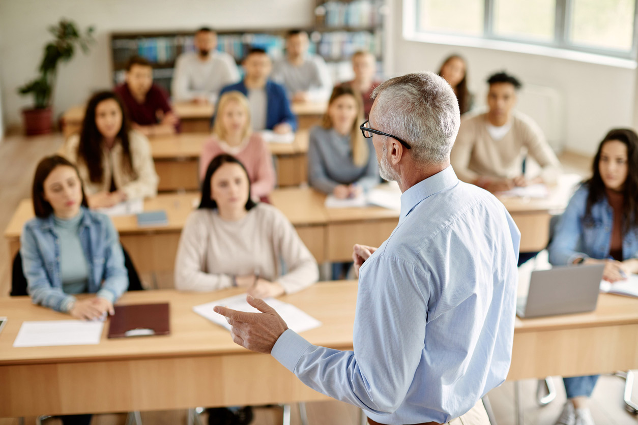 A teacher standing in front of a classroom, speaking to attentive students seated at desks with notebooks and laptops.