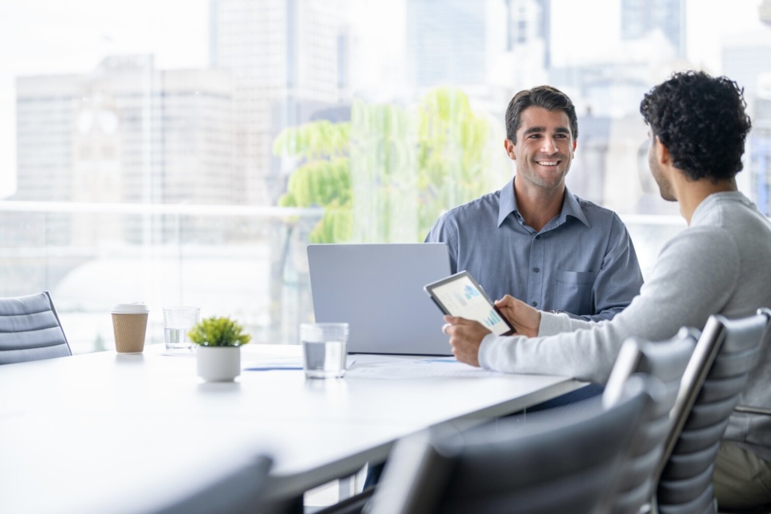 Two men sit in a bright white and grey office room and smile at one another.