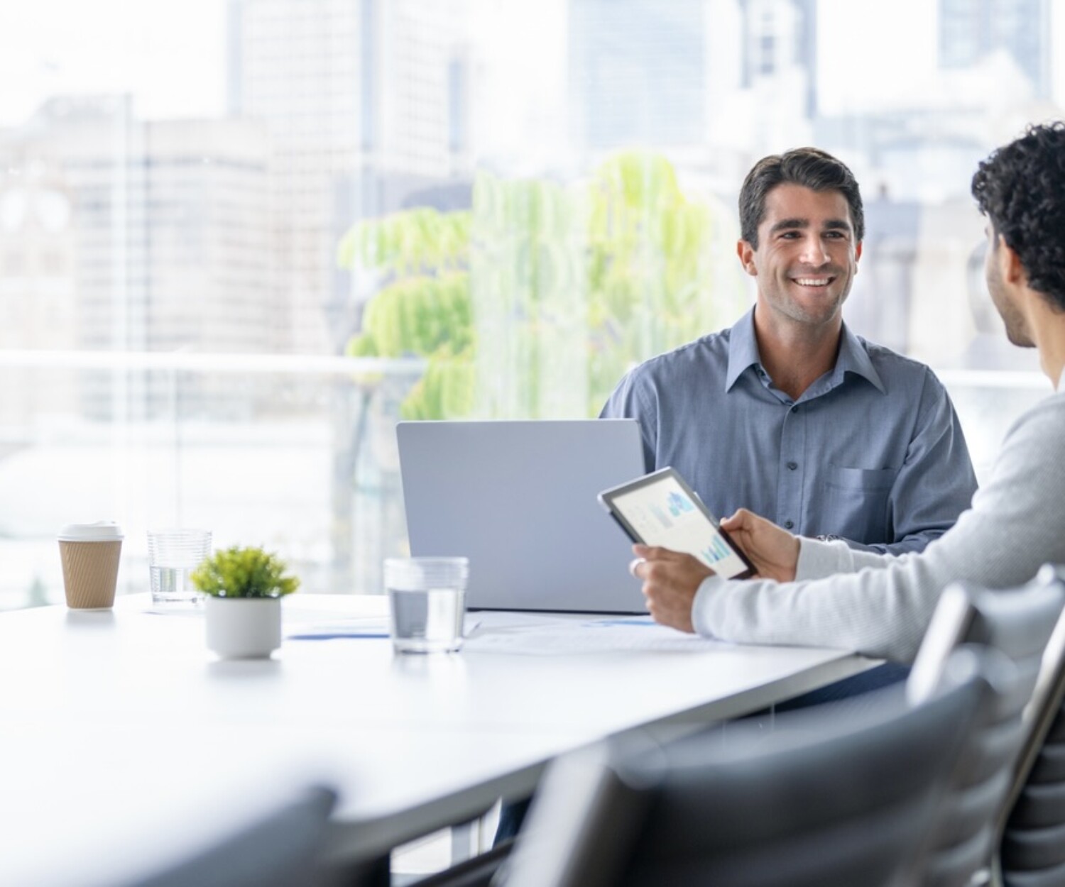 Two men sit in a bright white and grey office room and smile at one another.