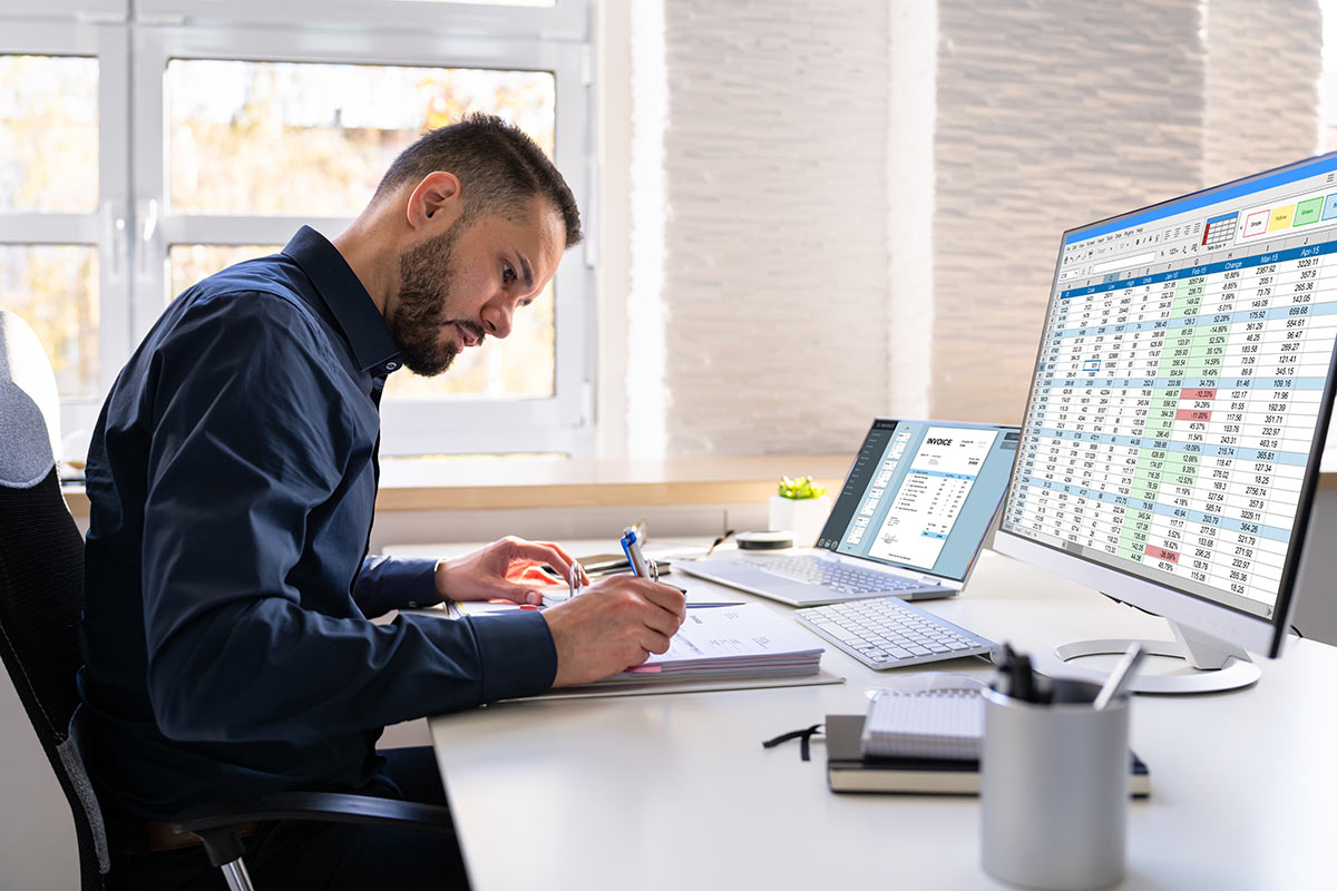 A person in a blue shirt writes in a notebook, surrounded by dual monitors displaying spreadsheets and documents in a bright office.