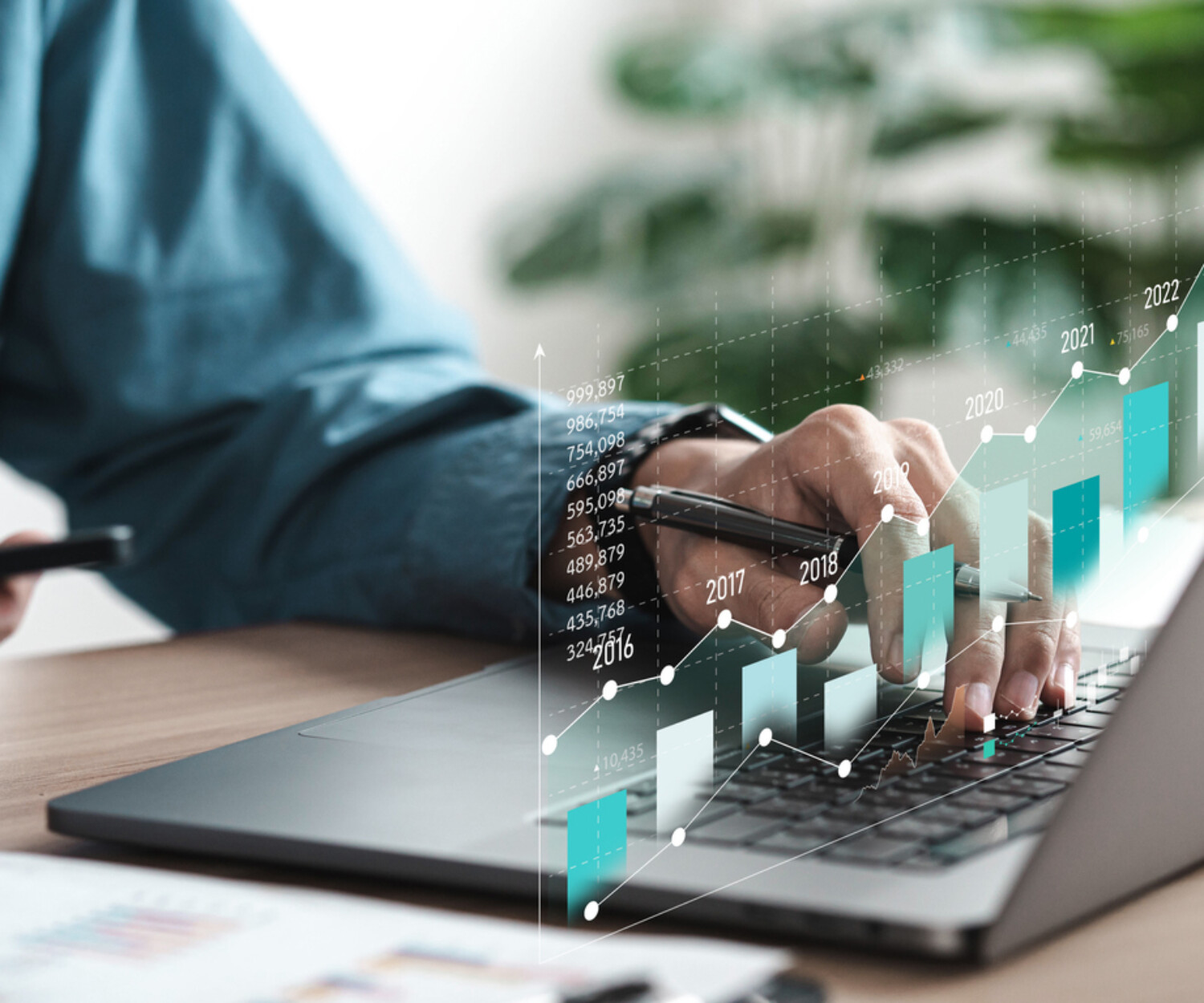 A person holding a pen types on a silver laptop. There is a bar chart overlaid on top of it